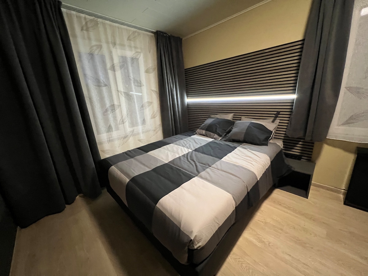 A spacious sleeping area features a bed with a striped black and grey duvet cover, complemented by two pillows. Nearby, floor-to-ceiling curtains frame a window, allowing natural light to filter in. The modern design includes a wooden accent wall behind the bed.