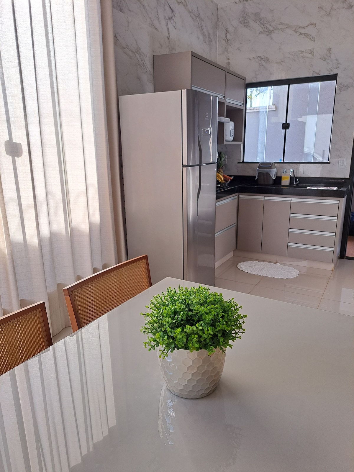 A sleek kitchen area is depicted, featuring modern cabinetry and stainless steel appliances. Natural light filters through sheer curtains, illuminating a dining table with a decorative plant in a textured pot, enhancing the inviting ambiance of the space.
