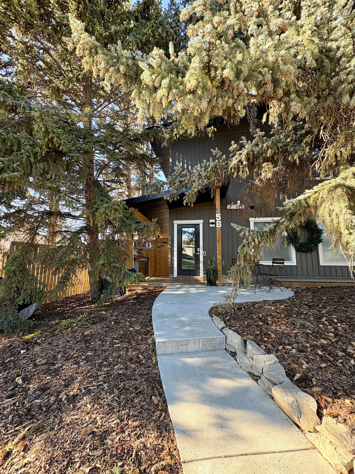 A welcoming entryway features a stone pathway winding through landscaped mulch, leading to a dark brown exterior with a glass door. Tall green trees provide shade while a small outdoor seating area is visible to the side, adding to the inviting atmosphere.