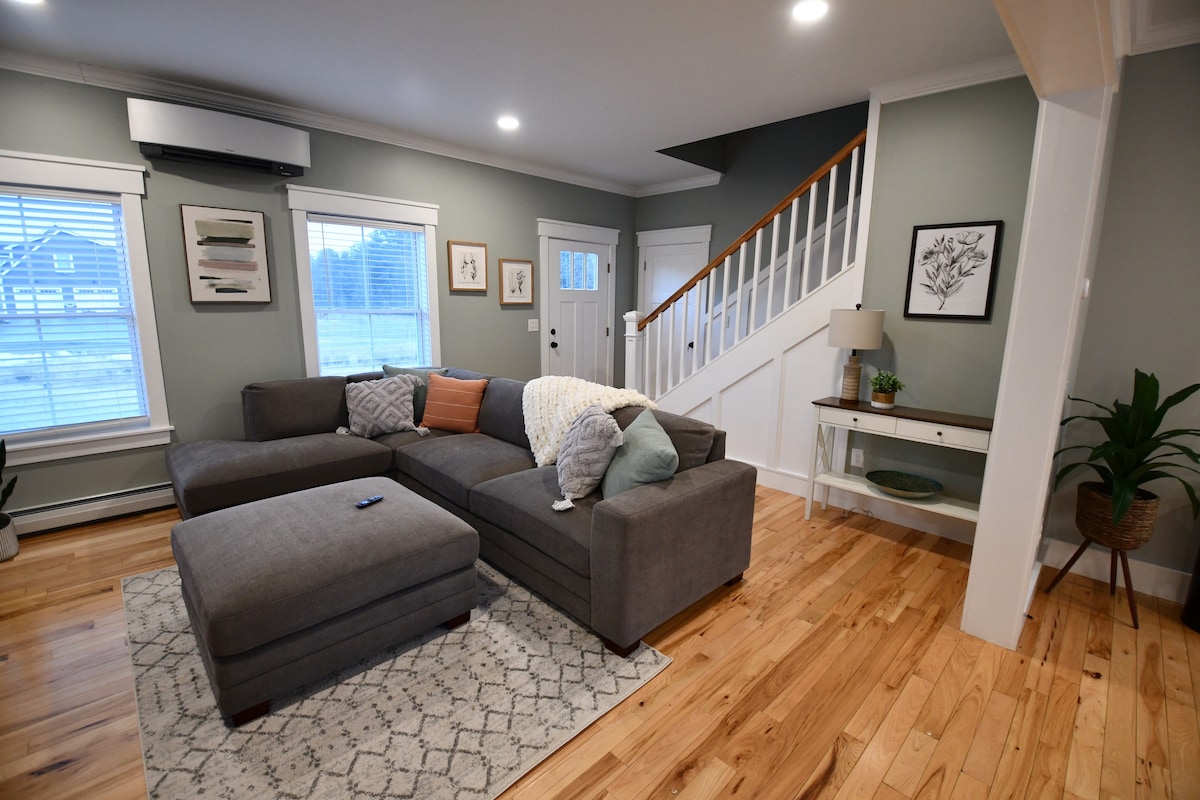 A spacious living area is highlighted by a large sectional sofa, accompanied by accent pillows. Natural light fills the room through multiple windows, with a patterned area rug underfoot. A small console table and green plant add touches of style to the inviting space.