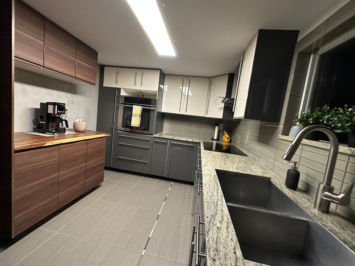 A modern kitchen features sleek cabinetry in contrasting tones of dark wood and light-colored finishes. The granite countertops offer ample workspace, complemented by stainless steel appliances. A double sink is visible, with a window above it allowing natural light into the space.