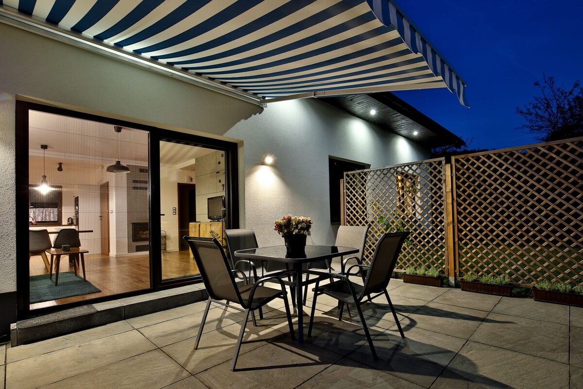 An outdoor patio is illuminated under a night sky, featuring a table surrounded by four chairs. A striped awning provides shade, while a lattice fence adds privacy. The view opens to a well-lit interior space, with visible elements of the modern home design.