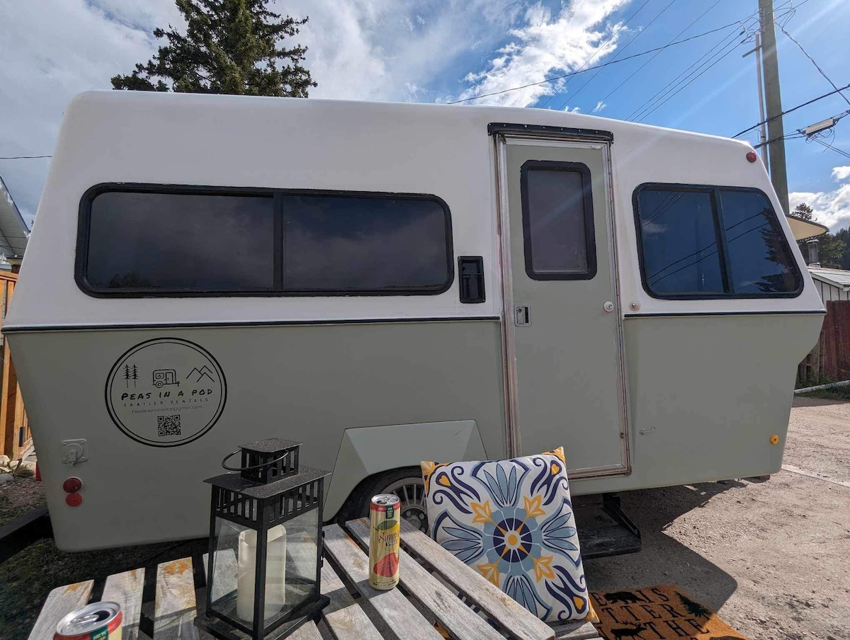 A vintage trailer is depicted, featuring a clean exterior with a soft green and white color scheme. Large windows enhance natural light, while a nearby table showcases a colorful patterned chair, a lantern, and beverages. The setting conveys a relaxed outdoor atmosphere.
