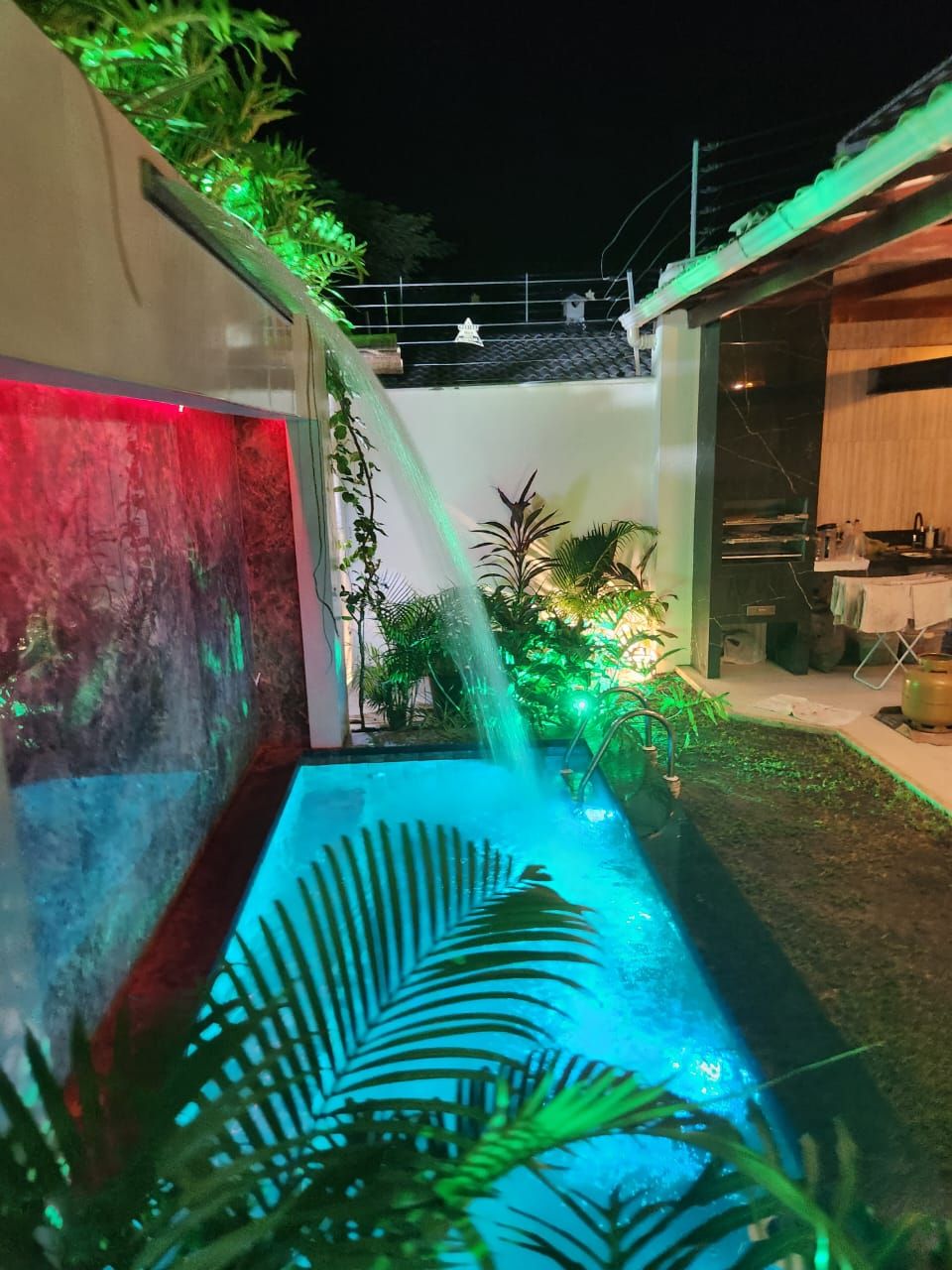 A serene swimming area is enhanced by soft lighting and a cascading water feature, surrounded by lush tropical plants. The pool's inviting blue water contrasts with the vibrant greens and ambient lights, creating a tranquil atmosphere at night.