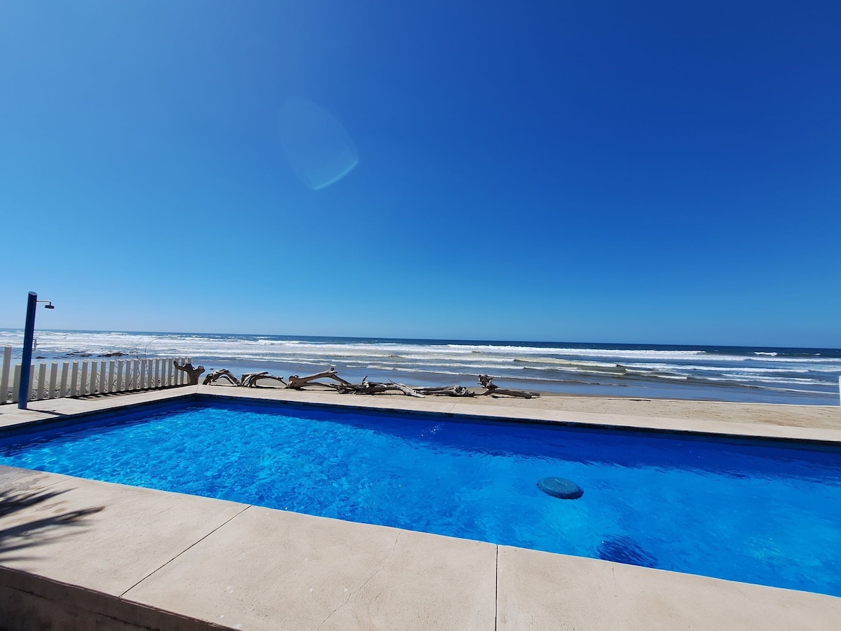 A clear blue swimming pool is positioned adjacent to the beach, with the ocean waves visible in the background. Sunlight reflects off the water's surface, creating a calm and inviting scene under a bright blue sky.