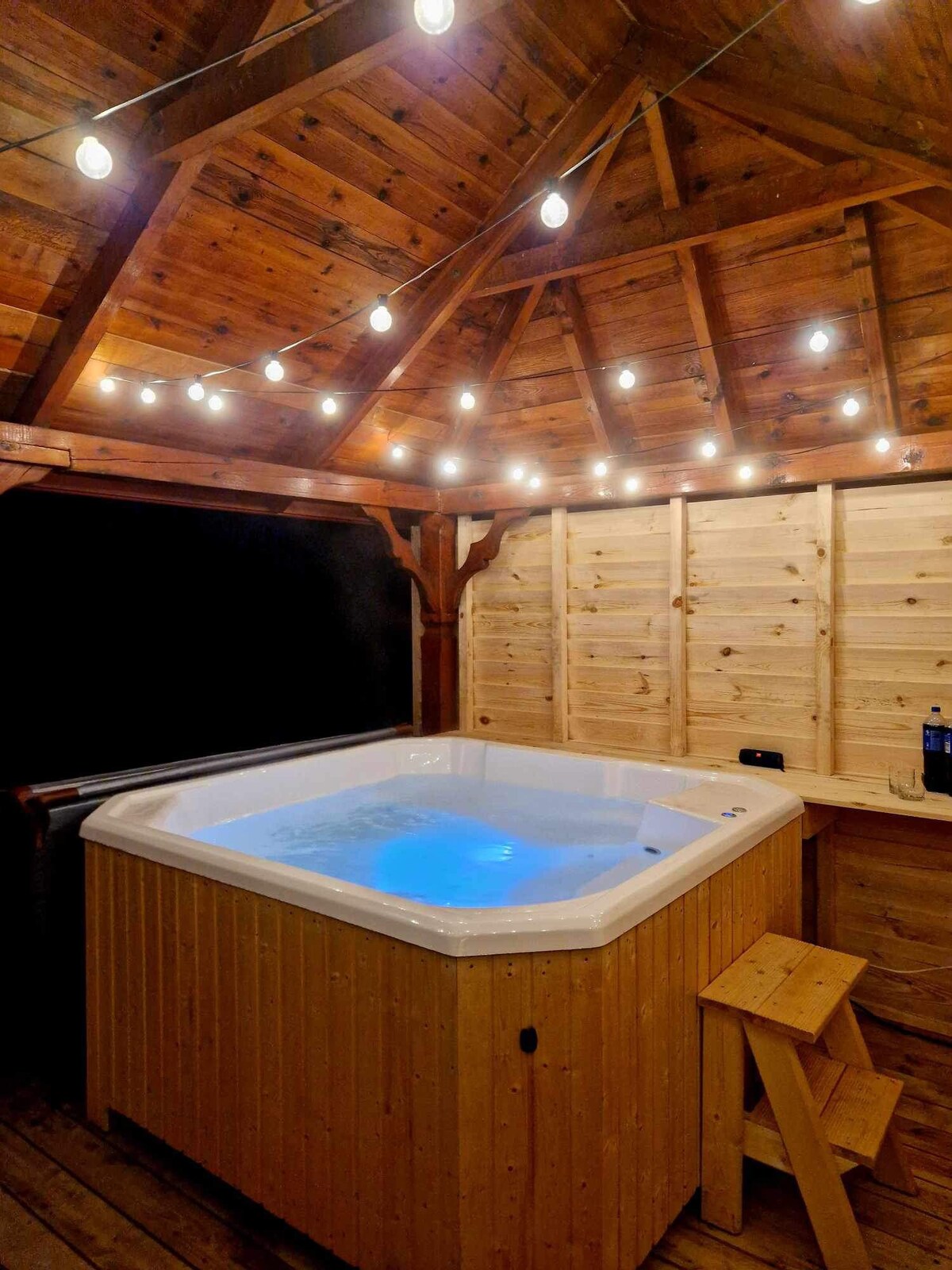 A hot tub with glowing blue water is nestled under a wooden gazebo, surrounded by warm string lights. The natural wood structure features exposed beams and paneled walls, enhancing a cozy, inviting space for relaxation.