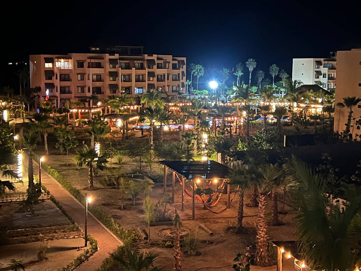 A vibrant outdoor space is illuminated by soft lights, showcasing palm trees and a well-maintained garden. Hammocks are placed under a shaded structure, inviting relaxation. The background features a two-story building with multiple balconies, contributing to a welcoming atmosphere.