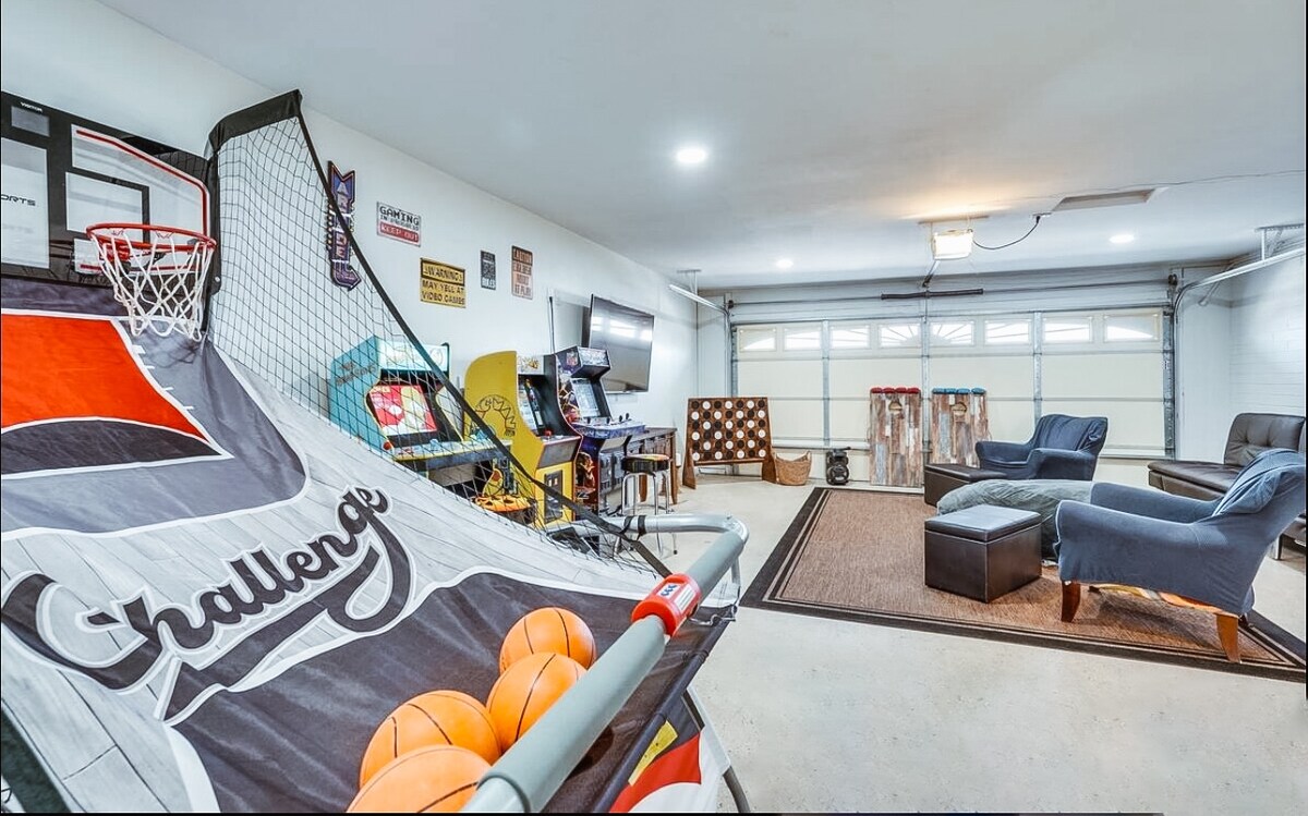 An entertainment room is equipped with a basketball arcade game and multiple classic arcade machines. Comfortable seating is arranged around a large rug, with ample natural light coming through the garage door. Vintage decor adds character to the spacious area.