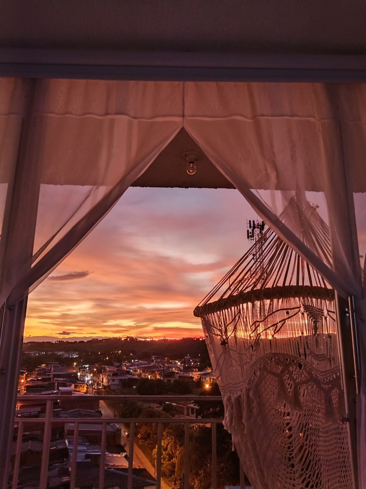 A view of a vibrant sunset is framed by sheer white curtains, highlighting orange and purple hues in the sky. Below, the rooftops of a lively neighborhood are visible, complemented by a macrame swing hanging from the balcony.
