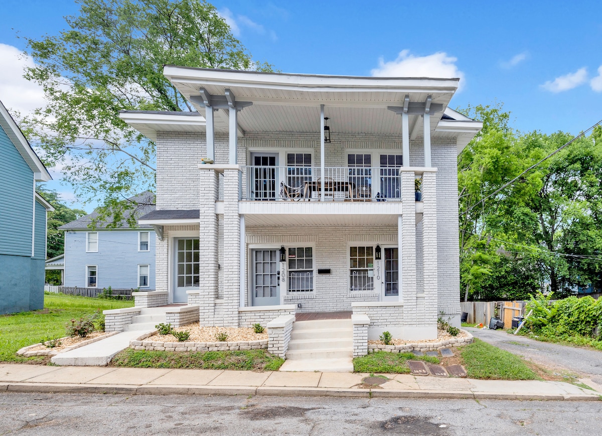 A charming two-story building features a light-colored brick facade and multiple front balconies. Symmetrical windows and front entrances are complemented by landscaped pathways leading to the building. Surrounding greenery adds a serene touch to the street view.