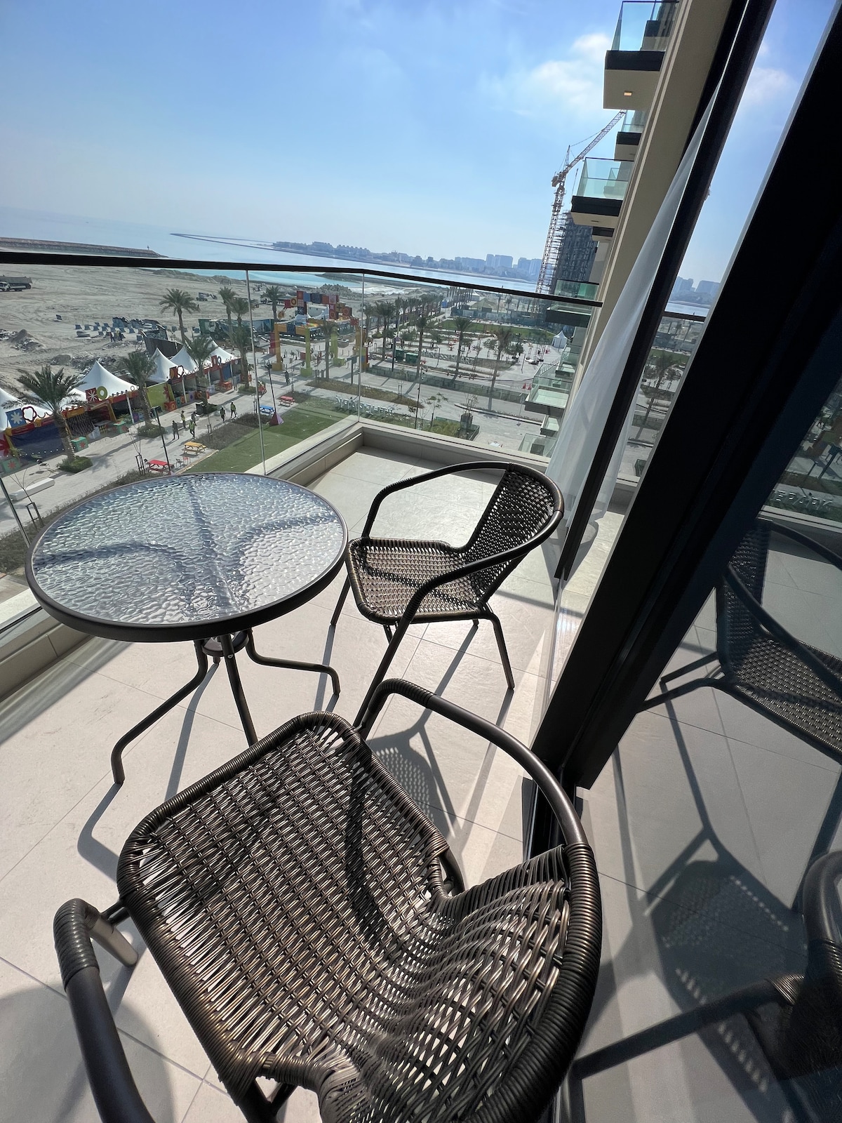 A balcony is seen featuring a round glass table surrounded by two woven chairs. The view includes a beach area and the distant shoreline, with palm trees lining the promenade. Bright daylight enhances the open space.