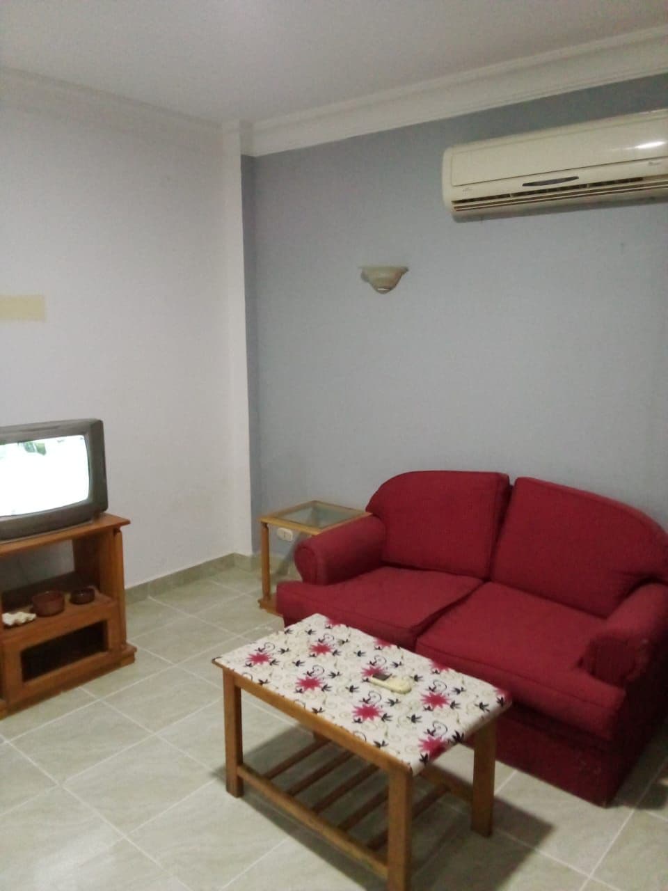 A compact living area is presented with a red sofa and a small coffee table, which is topped with a floral tablecloth. An air conditioning unit is mounted on the wall, and a television is placed on a wooden stand. Light-colored tiles cover the floor.