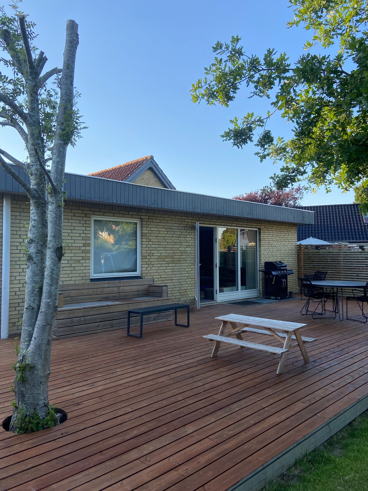 The outdoor terrace features a wood deck with a picnic table and a bench alongside a tree. Glass doors lead into the house, while a barbecue grill and a dining set are visible, providing options for outdoor meals and relaxation.