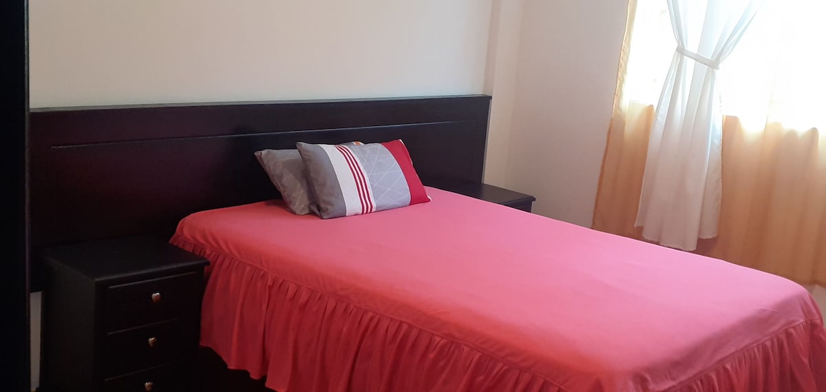 A bright sleeping area is presented with a spacious bed covered in a red bedspread. Two nightstands are positioned on each side, each featuring a drawer. Natural light enters through a window, which is dressed with light curtains, creating a cozy ambiance.