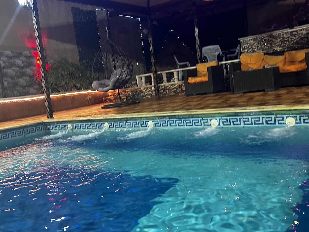 A serene pool area is presented, featuring clear blue water with gentle ripples. Comfortable seating is arranged nearby, including a hanging chair and lounge chairs adorned with bright cushions. Subtle lighting reflects off the water's surface, enhancing the tranquil environment.