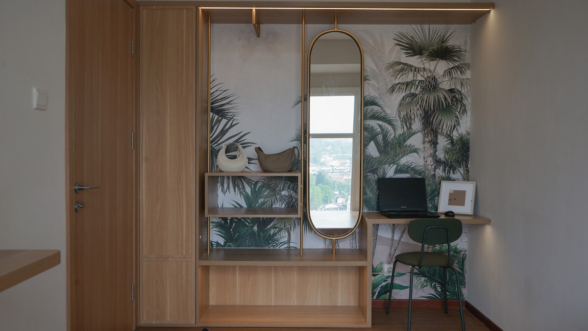 A functional workspace is featured with a desk and a comfortable chair, illuminated by natural light from a large window. The wall displays a vibrant mural of palm leaves, adding a touch of nature. Minimalist shelving holds decorative items and a mirror.