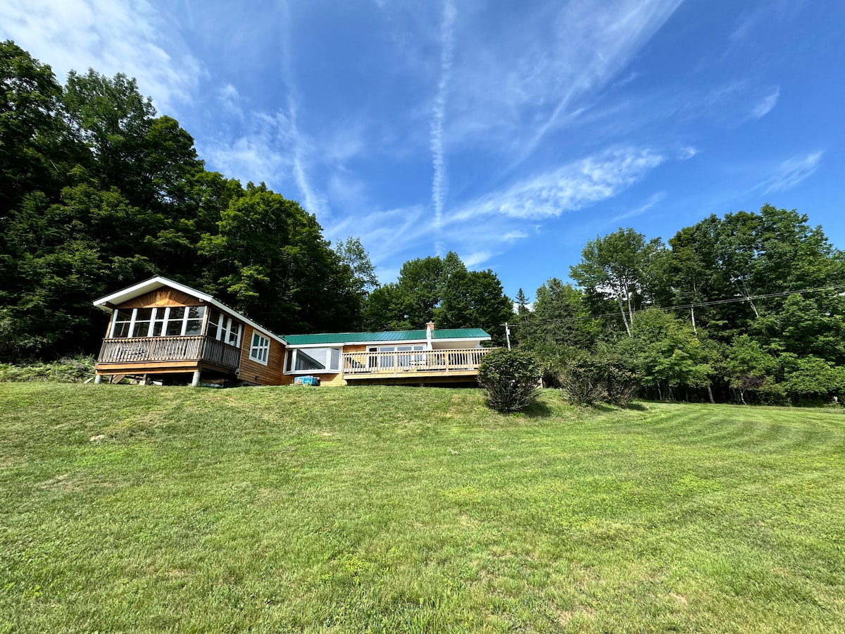 A cozy cabin is nestled on a grassy hillside, framed by tall trees. An expansive deck extends from the cabin, providing a vibrant outdoor space. The sky is clear with wispy clouds, enhancing the serene mountain retreat atmosphere.