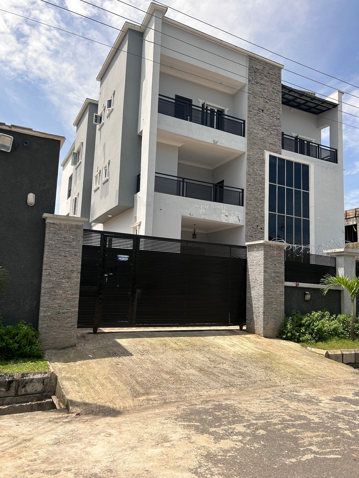 A modern three-story building is framed by a sleek black gate and driveway. The façade features a mix of textured stone and smooth white surfaces, with large glass windows allowing natural light to fill the interior.
