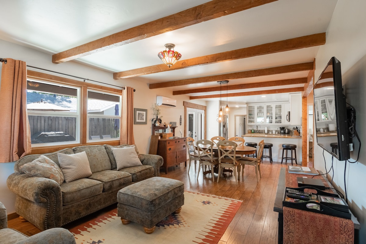 An open concept living area features exposed wooden beams and large windows allowing natural light. A cozy sofa is positioned near a TV, while a dining table with multiple chairs is seen. The updated kitchen is visible in the background, enhancing the inviting space.