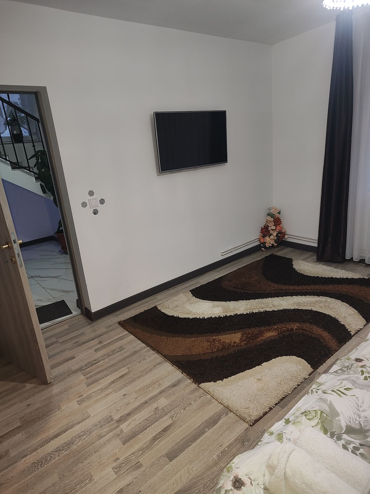 A clean and modern room features a wall-mounted television and a patterned area rug with warm tones. Natural light enters through a nearby window, complemented by dark curtains. Decorative items can be seen alongside the staircase in the background.