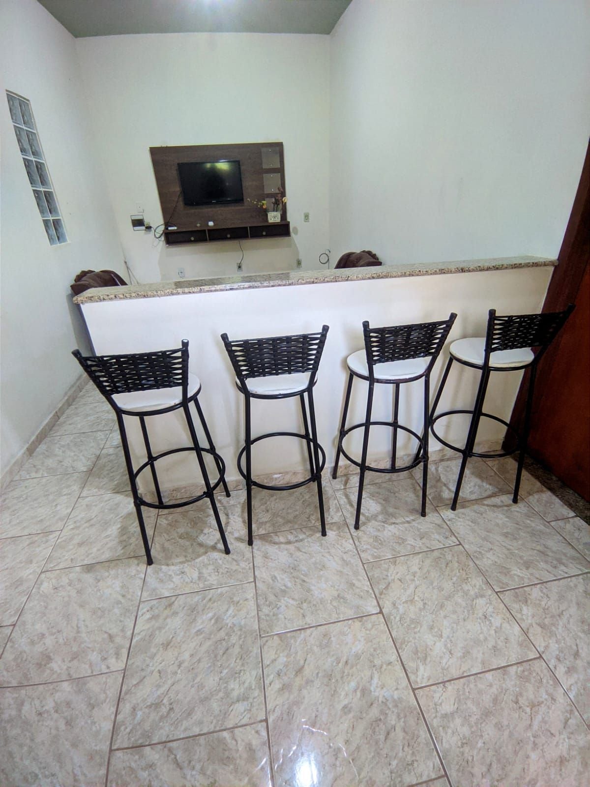 Four high-backed stools are arranged around a granite countertop. The tiled floor complements the neutral color scheme of the walls, while a mounted television and wall unit is visible in the background.