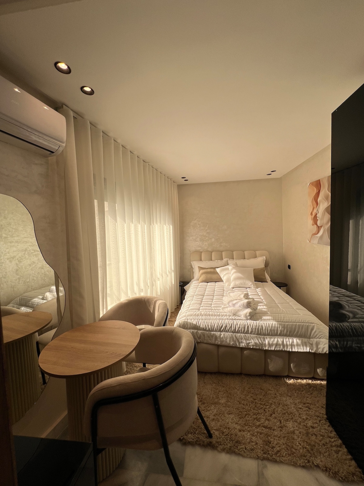 A cozy bedroom is shown with a spacious bed dressed in white linens. Natural light filters through sheer curtains, illuminating the room. A small round table and two upholstered chairs complement the decor, and a mirror adds depth to the space.