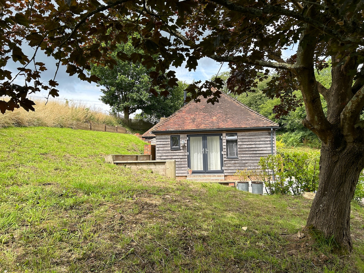 A charming self-contained annex is situated at the edge of a grassy hill, surrounded by trees. The exterior features a wooden façade and double doors that lead to a private patio. A well-maintained landscape adds to the serene atmosphere.