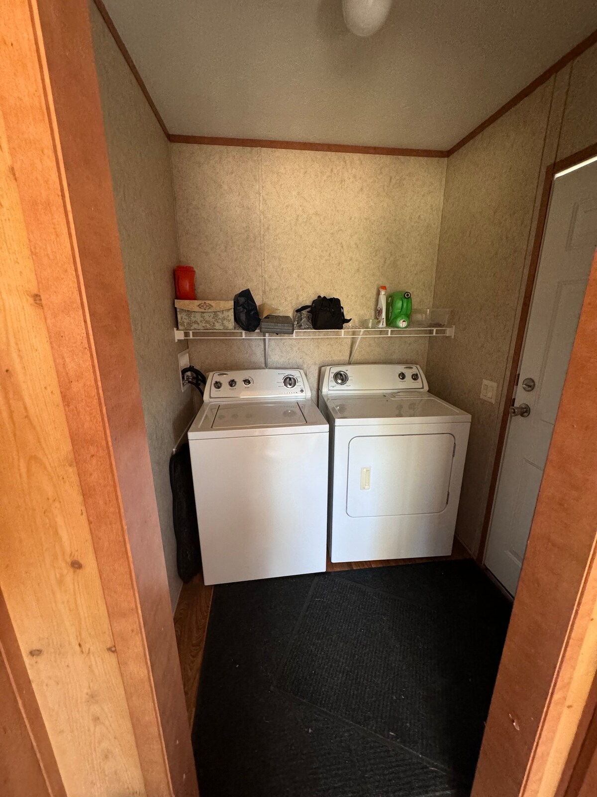 A laundry area features two white appliances, a washer and dryer, positioned side by side. Shelves above hold various items, including a few containers. The floor is covered with a black mat, contributing to a functional and tidy space.