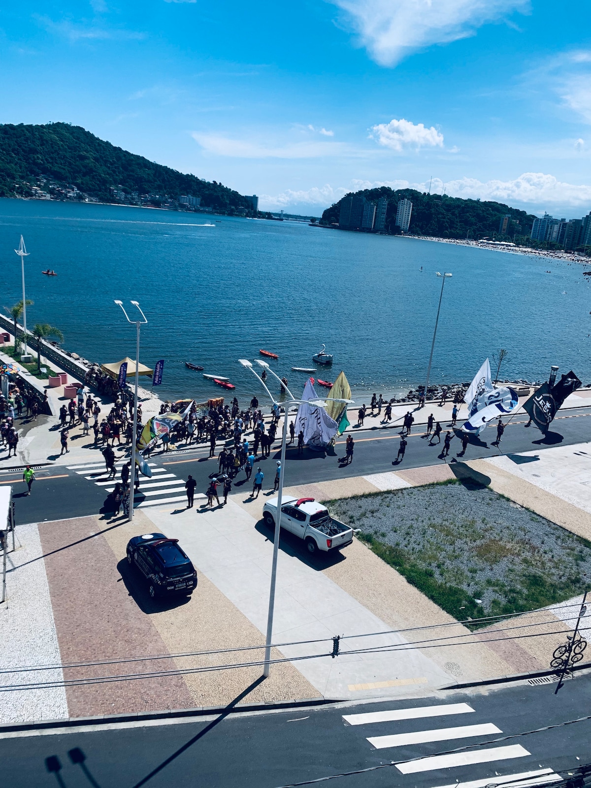 A view of a busy waterfront area captures a crowd participating in an event along the promenade. Flags and banners are displayed, while boats can be seen on the water. The skyline is defined by buildings atop a lush green hill in the background.