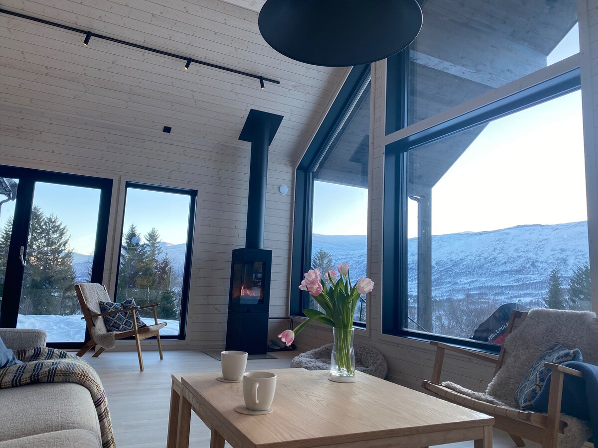A cozy living area features large windows that provide scenic mountain views. A modern wood stove sits in the corner, while a wooden table is set with two white cups. Fresh flowers are displayed in a vase, adding a touch of color to the space.