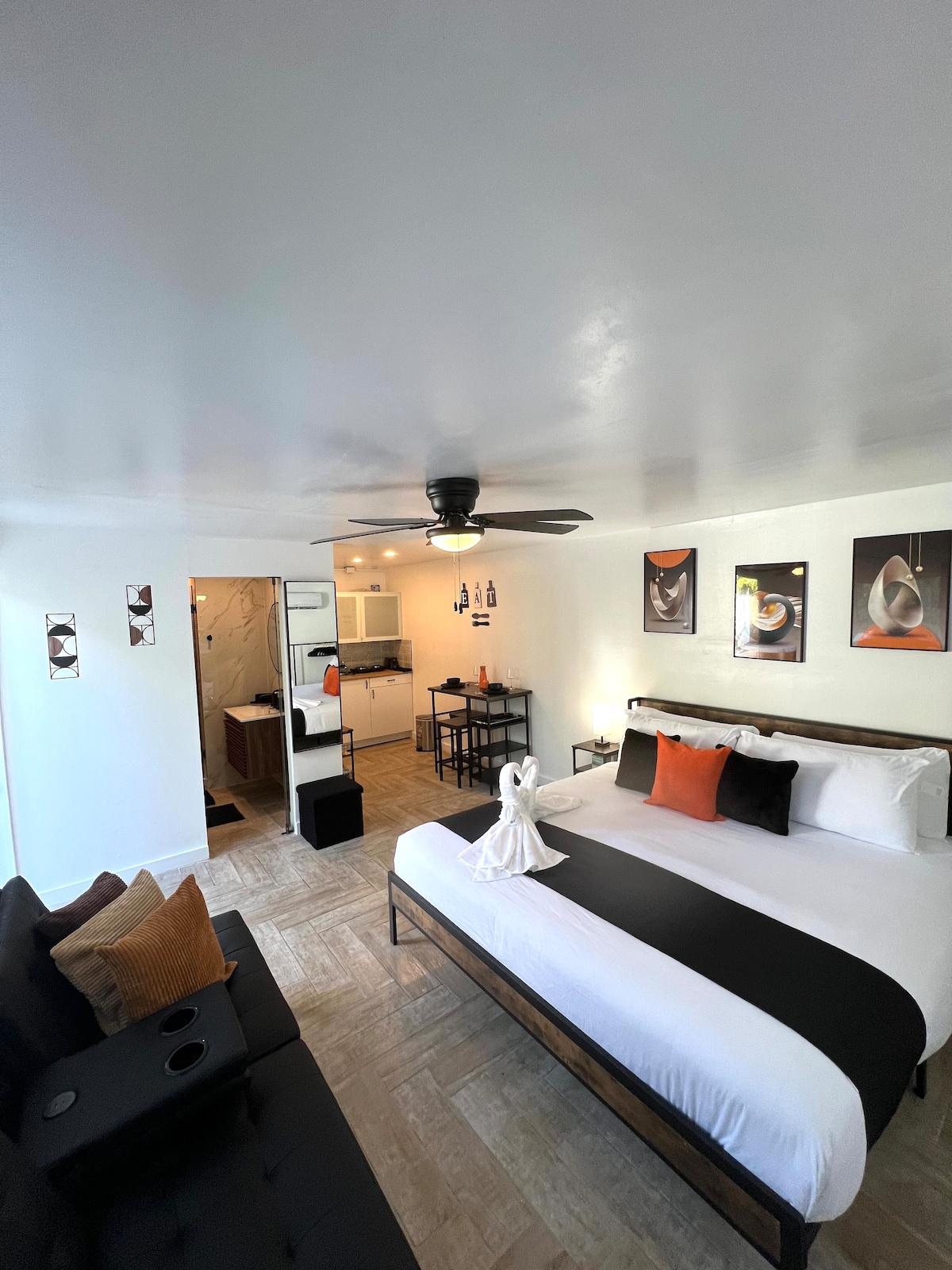 A modern studio space is showcased, featuring a spacious king bed with black and white bedding accented by throw pillows in orange. A comfortable seating arrangement is visible, and artwork adorns the walls. The open layout connects to a kitchenette area at the back.