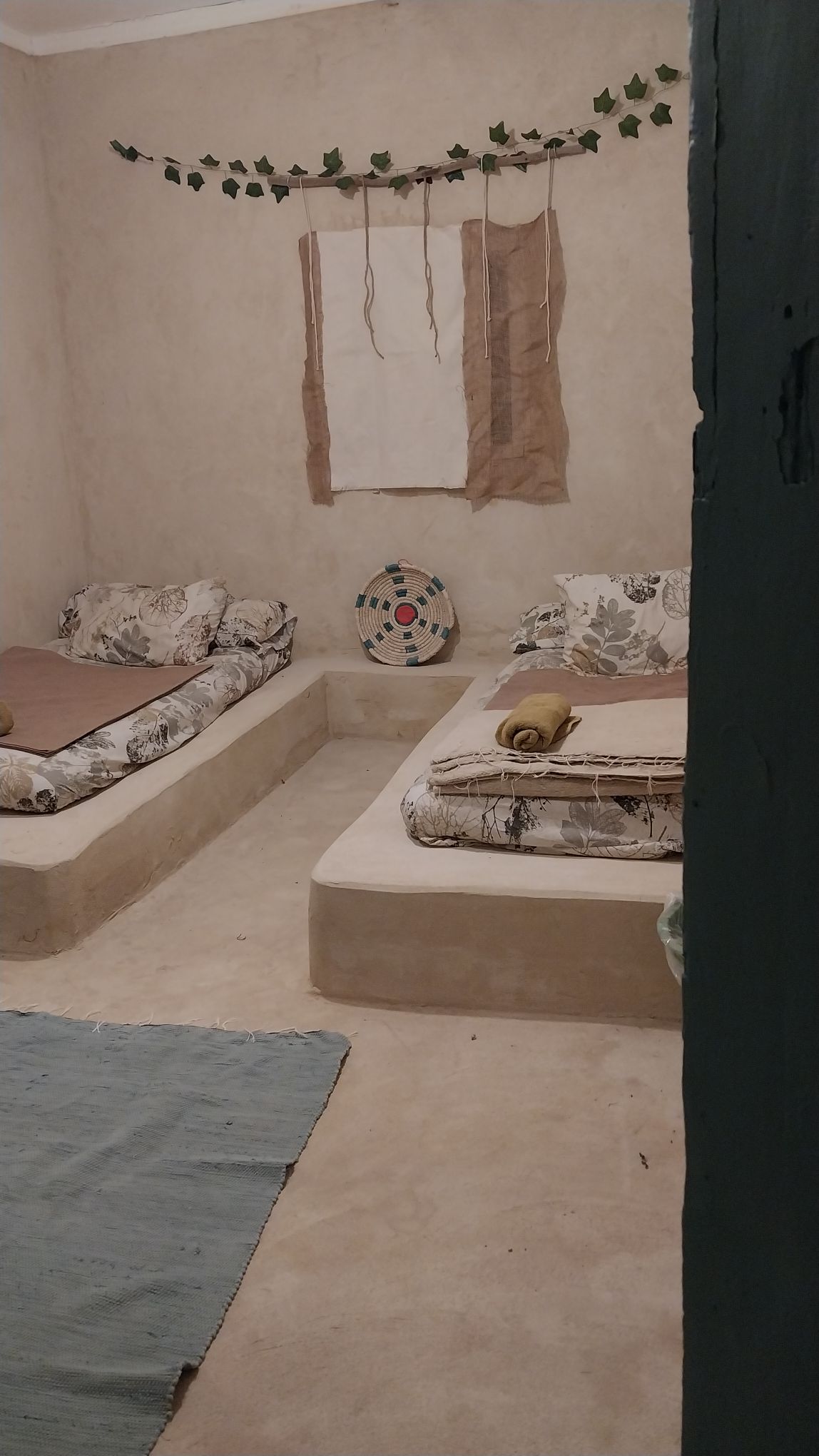 A serene sleeping area features two low-profile beds adorned with floral bedding. Natural tones create a calming environment, while minimal decor, including greenery and a circular wall art piece, adds subtle character. Light filters in through a simple window, enhancing the peaceful ambiance.