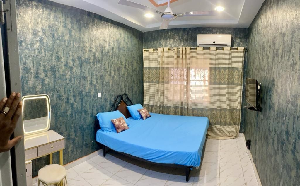A spacious sleeping area features a large bed dressed in blue linens, complemented by decorative pillows. A vanity table is positioned nearby, and a wall-mounted television is visible. Natural light filters through the partially closed curtain, enhancing the room’s tranquil ambiance.