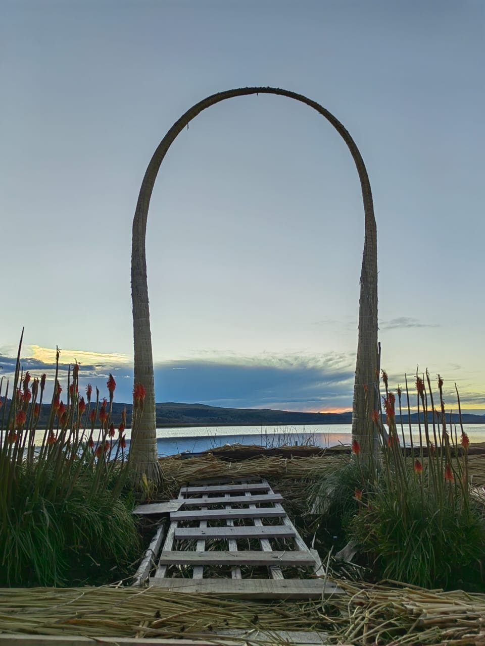 A wooden pathway leads towards a large archway made of natural materials, framed by lush greenery and tall plants with red flowers. The serene lake and distant hills are visible in the background, bathed in soft evening light.