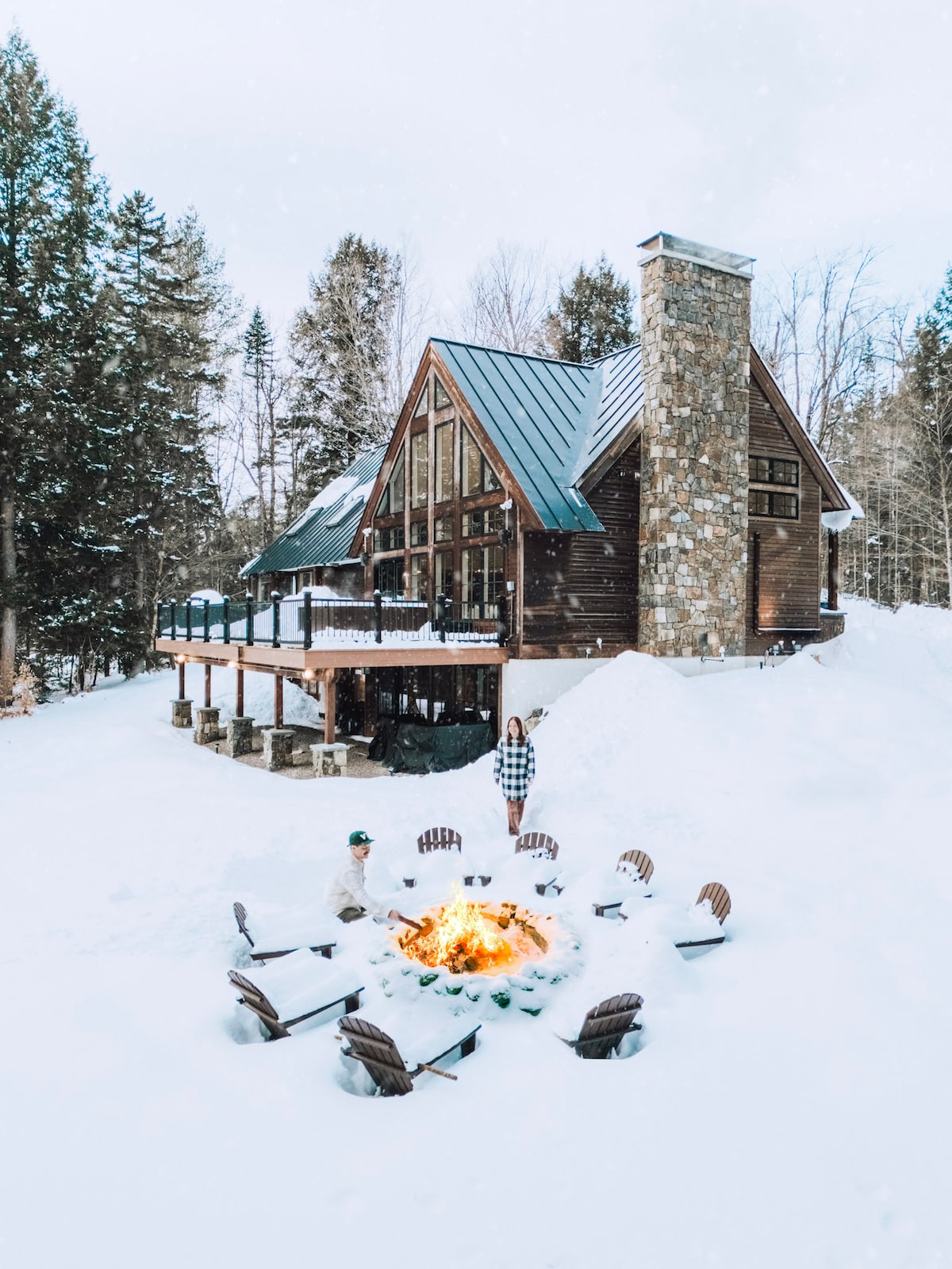 A large ski lodge features a stone chimney and sloped metal roof, surrounded by snow-covered trees. A circular fire pit with multiple chairs is situated in the foreground, inviting relaxation amidst a winter landscape.