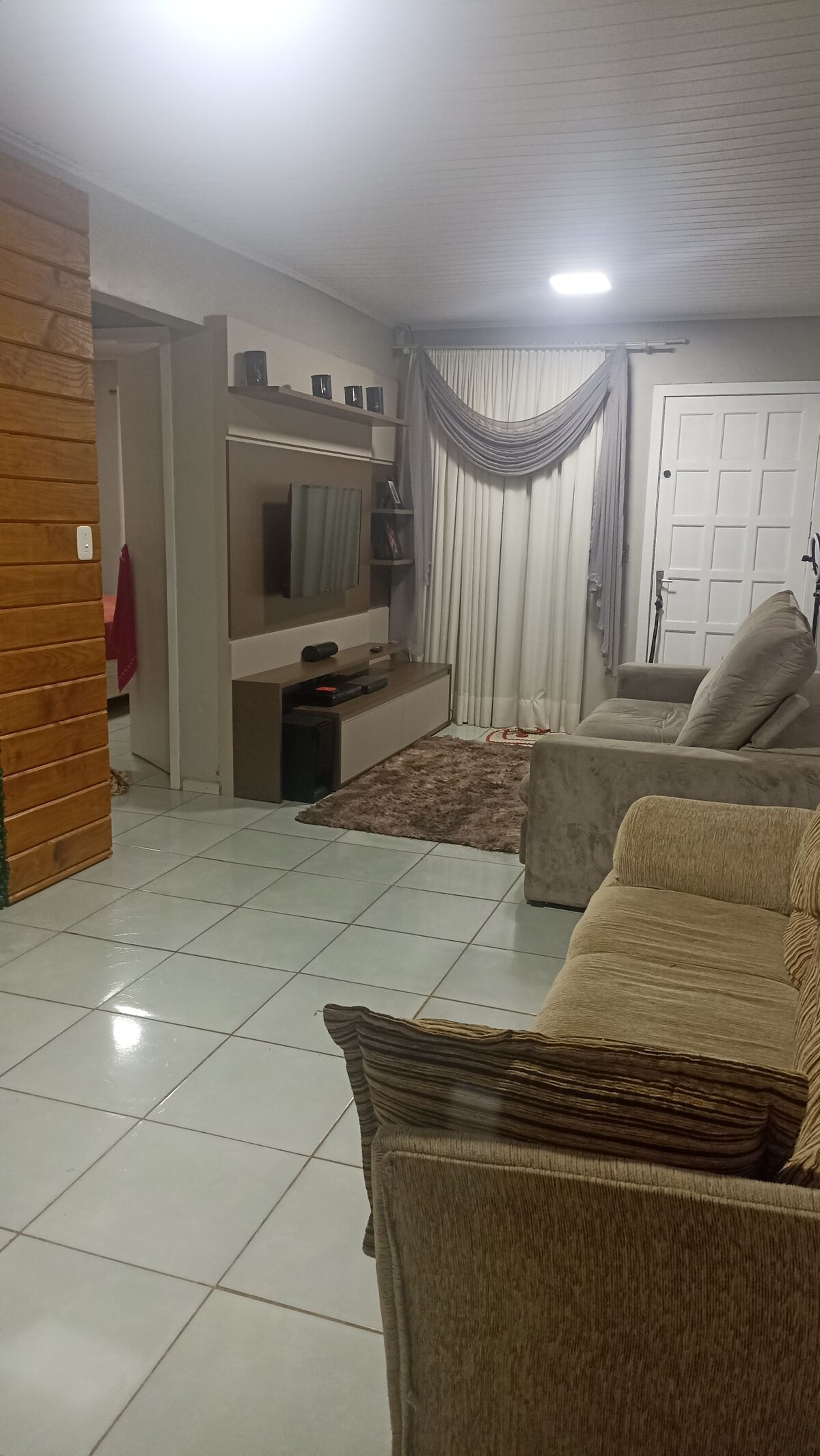 A comfortable living area features two large sofas facing a flat-screen TV mounted on a light-colored wall. A softly patterned rug lies beneath, and natural light filters through sheer curtains. Nearby, a door with paneling provides access to an entrance.