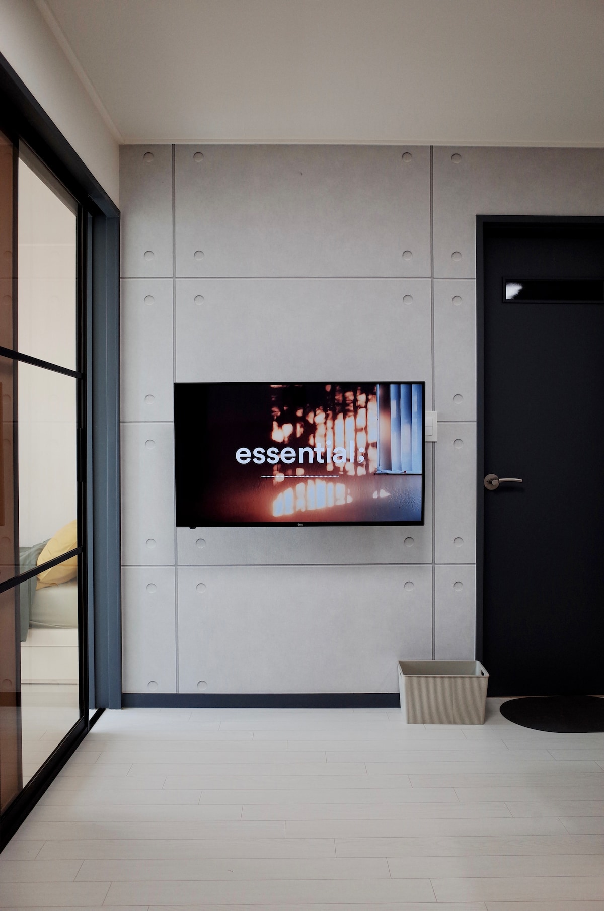 A modern wall is equipped with a flat-screen television displaying an abstract image. A sleek door is visible nearby, along with a minimalist waste basket on the floor. The flooring features light-colored planks, enhancing the contemporary atmosphere.