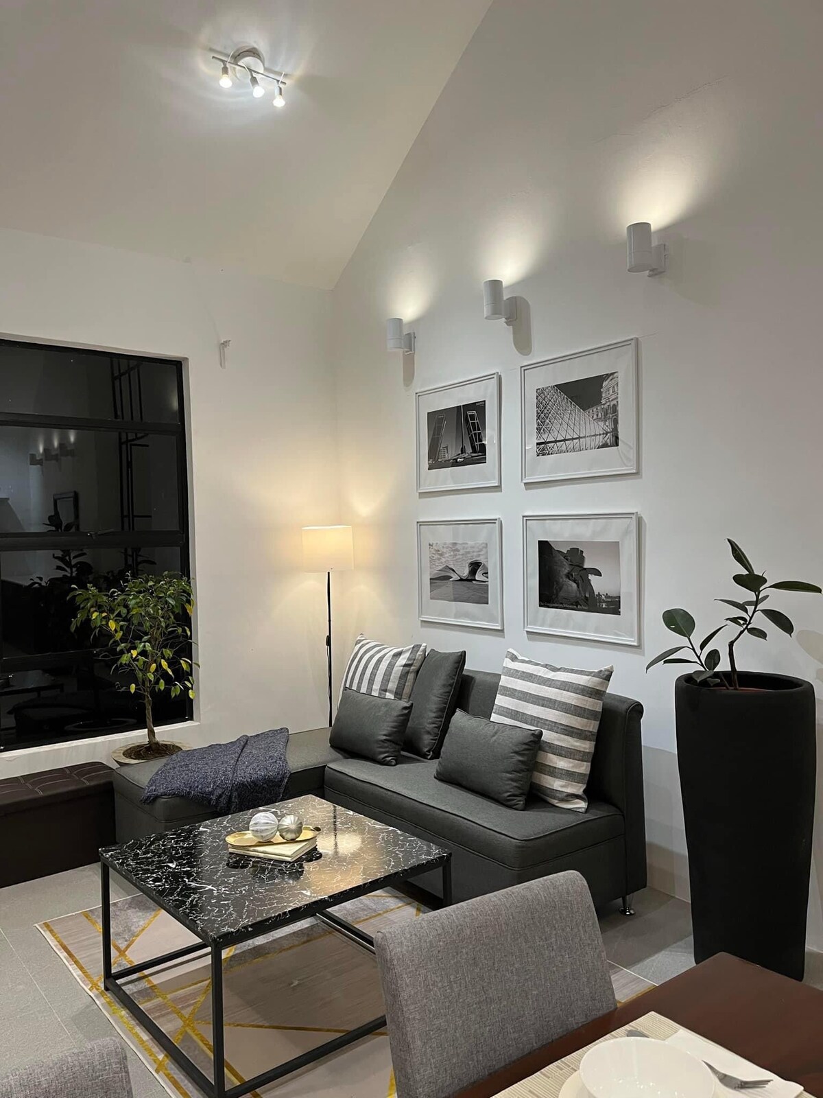A modern lounge area is depicted with a sleek grey sofa adorned with striped pillows. A glass coffee table sits on a textured rug, while framed black-and-white photographs are arranged above. Soft lighting enhances the ambiance, and potted plants add a touch of greenery.