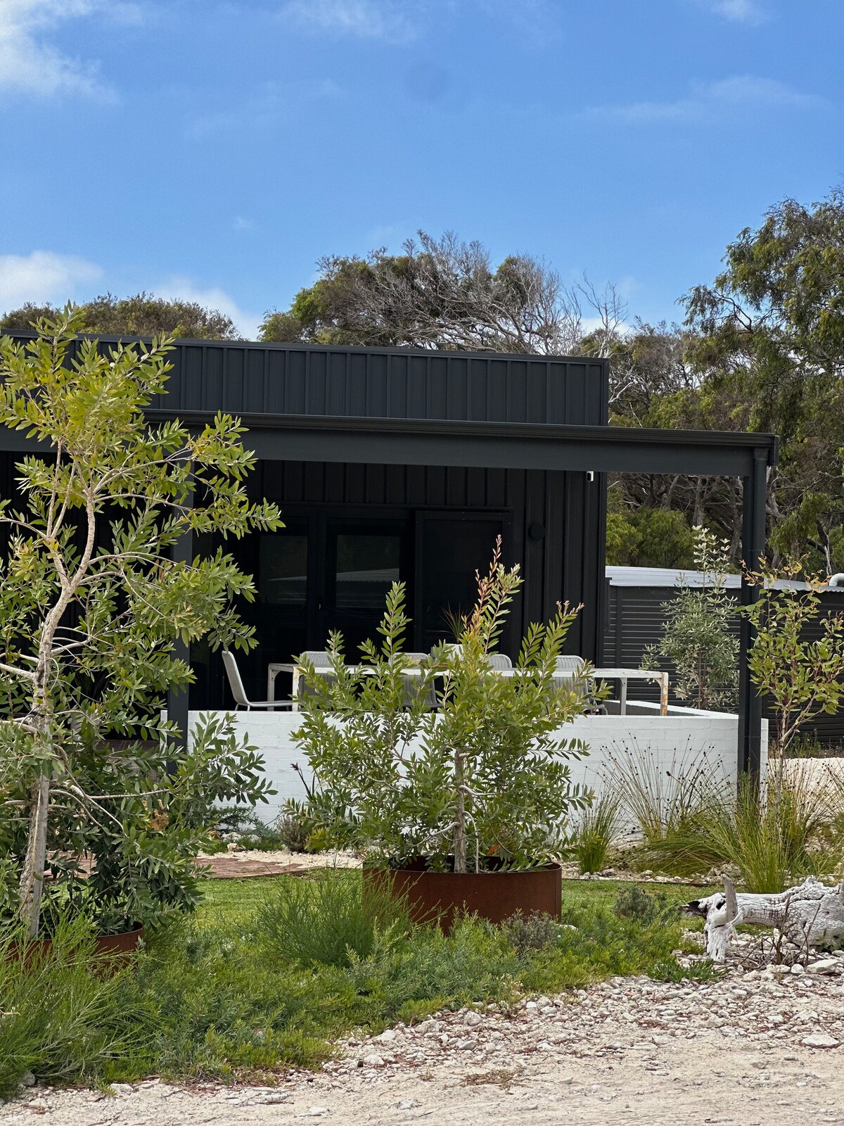 The exterior of the accommodation features a modern black structure with large windows, surrounded by lush native landscaping. A spacious deck with seating is visible, inviting guests to enjoy the natural setting. A rustic planter box enhances the aesthetic, contributing to the peaceful atmosphere.
