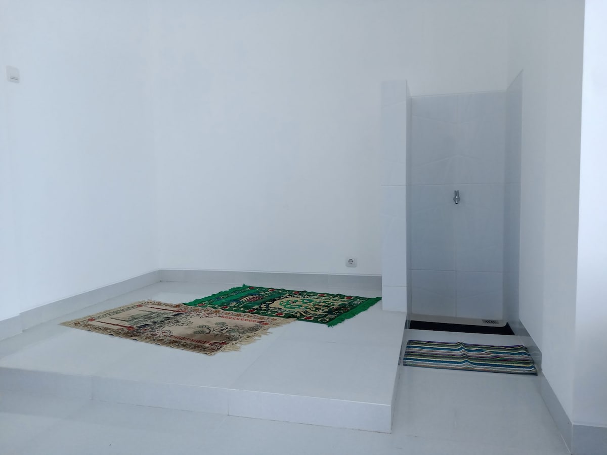 The room features a clean and minimalistic design with white walls and flooring. Two rugs are placed on the floor. A shower area is present against one wall, offering a simple yet functional layout.