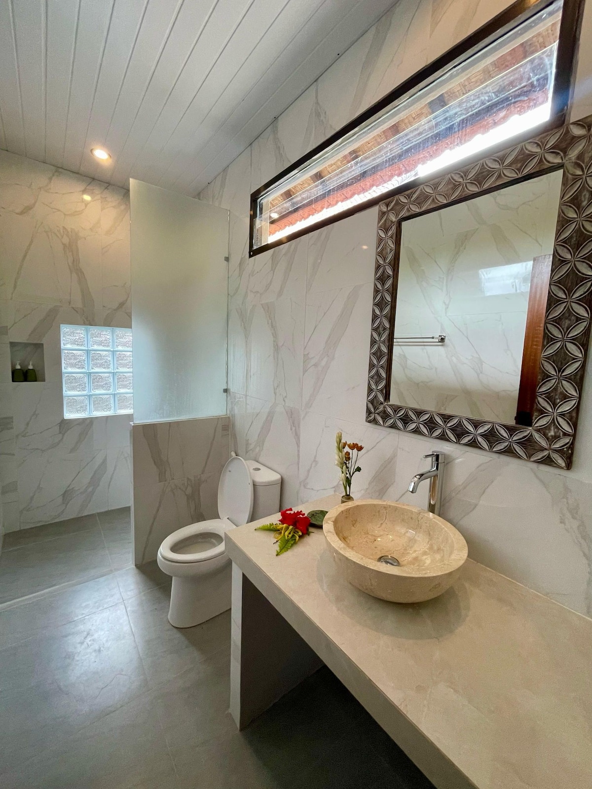 A modern bathroom features elegant marble walls and a sleek stone sink. A wooden-framed mirror adds a touch of warmth. Natural light filters through a small window, illuminating the clean and uncluttered space with a small arrangement of flowers placed on the countertop.