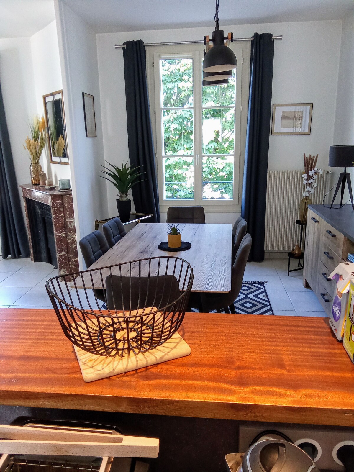 The dining area features a wooden table surrounded by upholstered chairs, positioned near a large window framed by dark curtains. A decorative basket is placed on the countertop in the foreground, with a stylish light fixture hanging overhead, adding to the room's modern aesthetic.