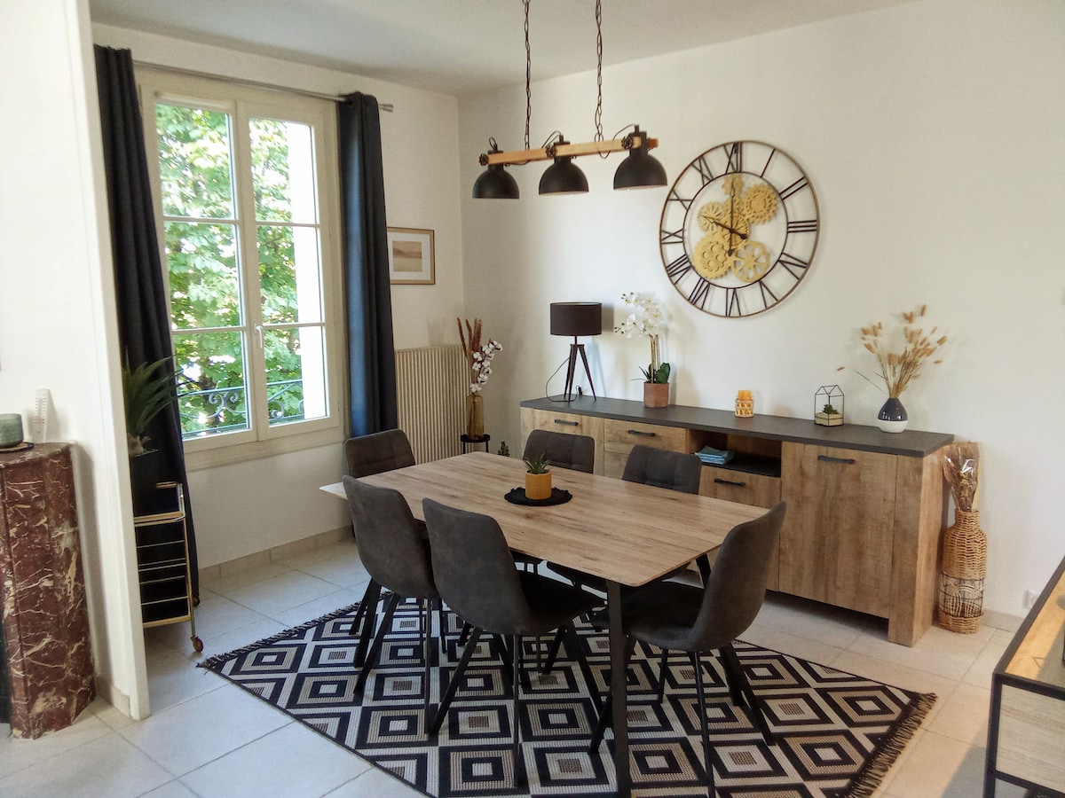 The dining area features a wooden table surrounded by six upholstered chairs. A patterned rug lies beneath, while a large clock and decorative elements adorn the walls. Light enters through the tall windows, framed by dark curtains, enhancing the space's openness.