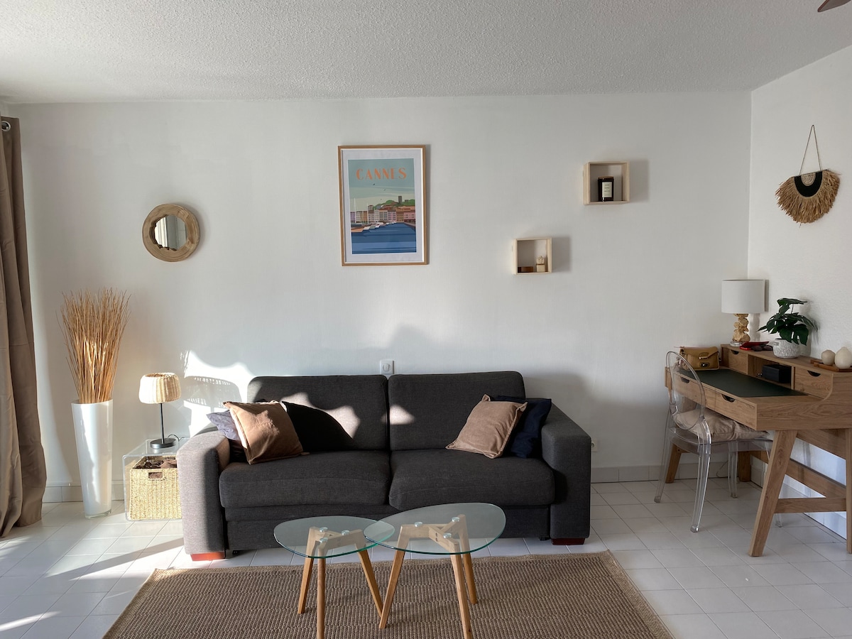A bright living area features a gray sofa with decorative pillows, paired with a glass coffee table. A wooden desk is arranged nearby, and art and decorative items adorn the walls. Natural light filters in through window curtains, complemented by a woven plant display.