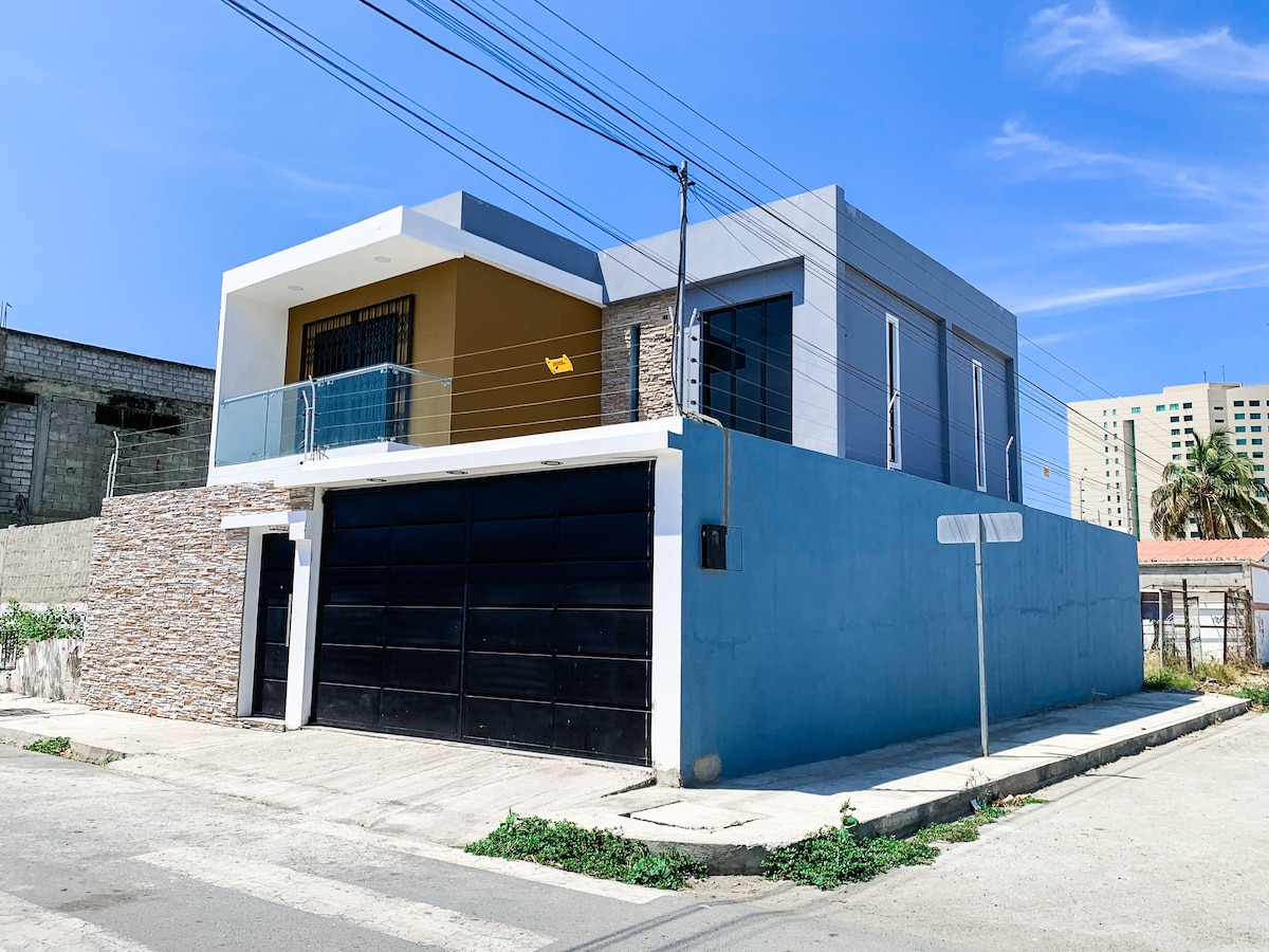 A modern two-story house is presented with a stylish facade, featuring textured stone and smooth blue walls. Large windows invite natural light, while a spacious driveway with a sleek black garage door is visible in the foreground.