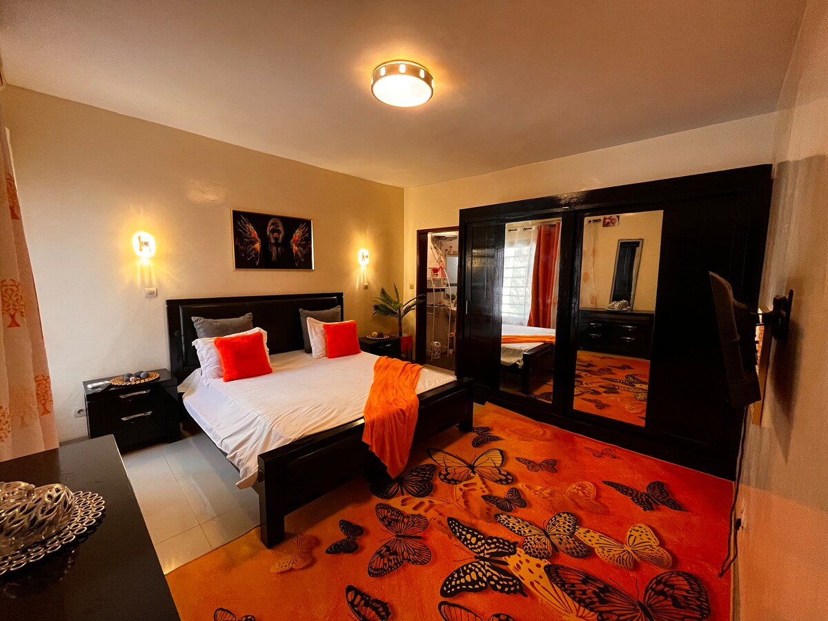The bedroom features a spacious bed adorned with orange and white accents, complemented by a large mirror and vibrant butterfly-patterned carpet. Natural light filters through the window, creating a warm ambiance within the elegantly designed space.