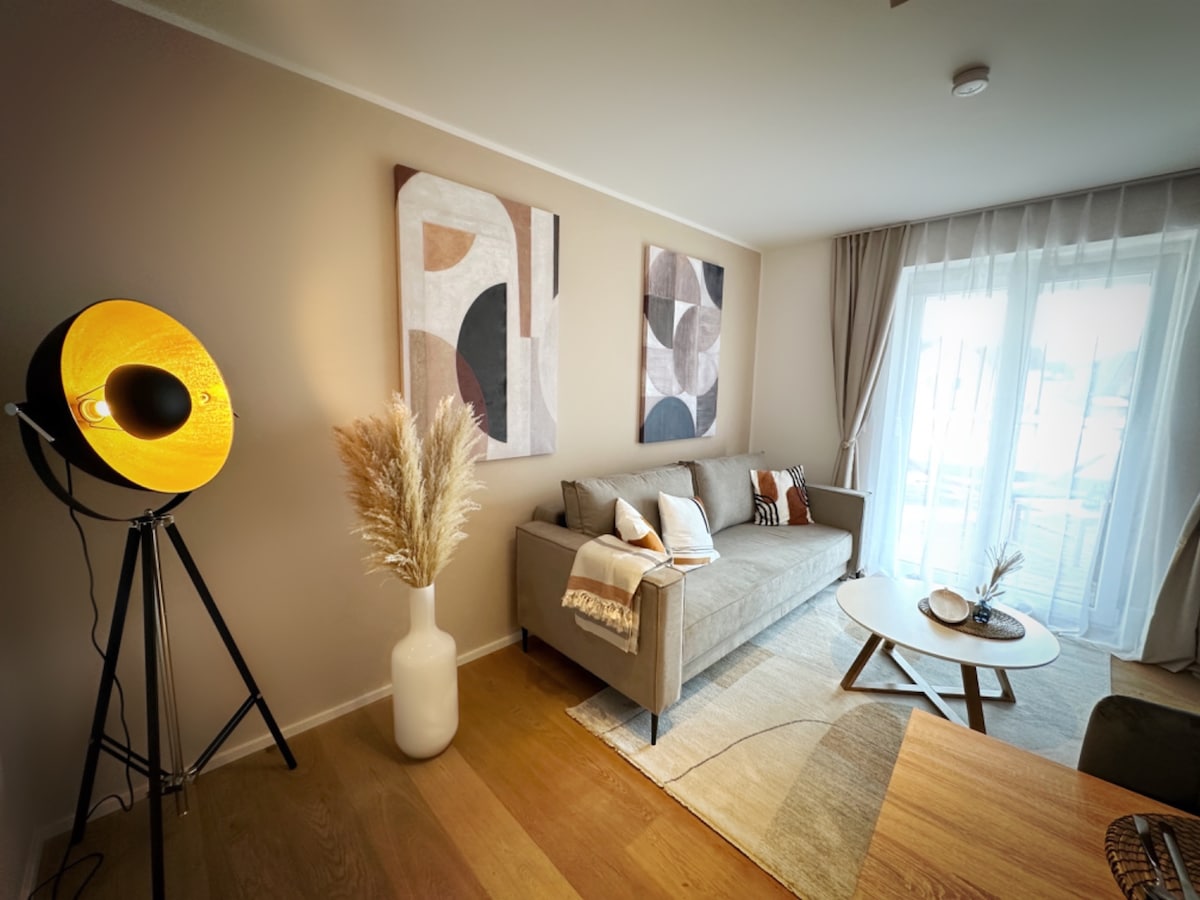 A modern living space is presented, featuring a comfortable sofa adorned with decorative cushions. An artistic lamp stands beside the sofa, illuminating the area. Neutral-toned artwork decorates the walls, and a round coffee table is positioned at the center, enhancing the inviting ambiance.