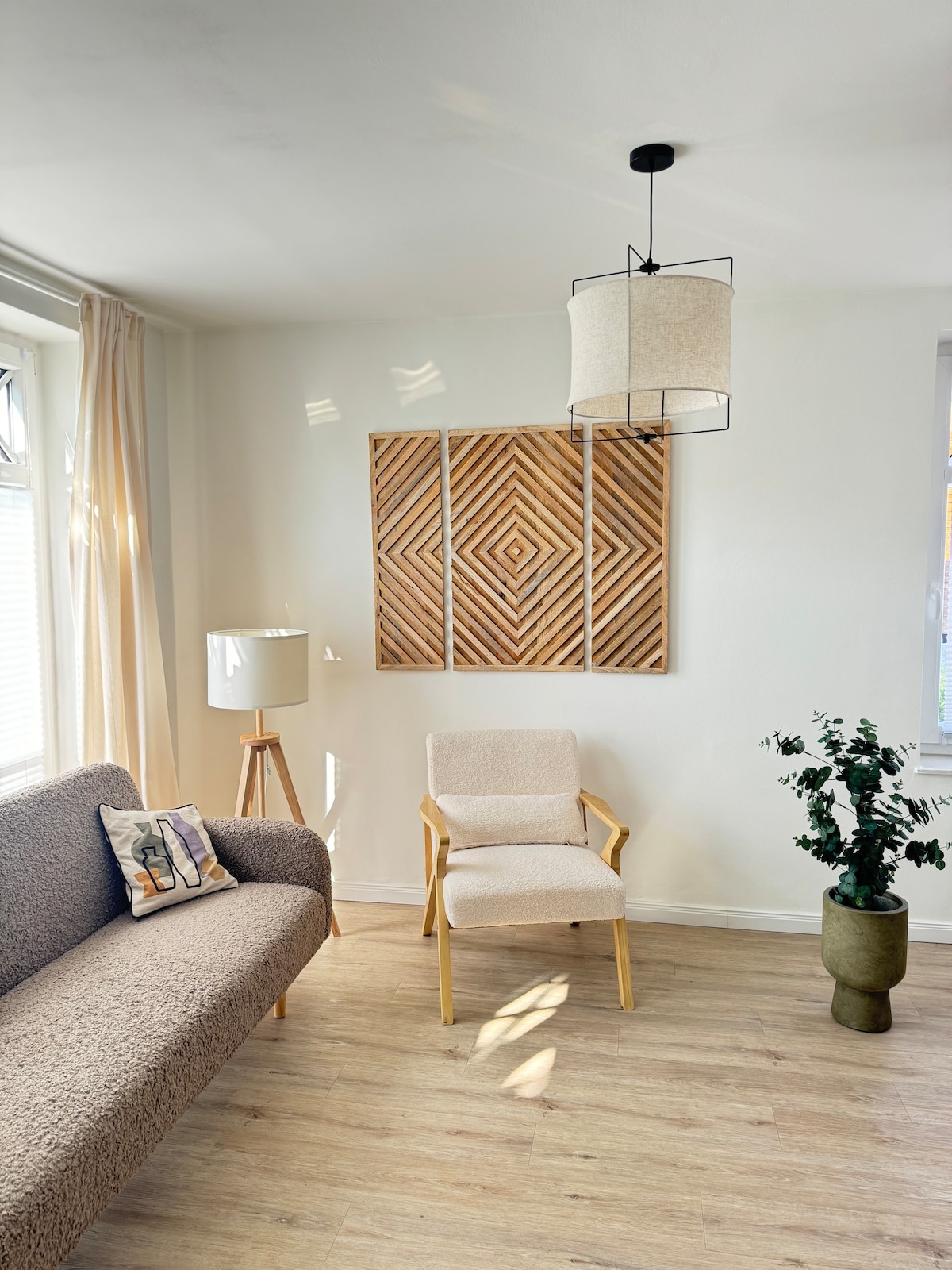 A bright living area is presented, featuring a textured sofa paired with a light-colored armchair. A wooden wall art piece enhances the space, while a potted plant adds a touch of greenery. Soft sunlight filters through nearby windows, creating a calm ambiance.