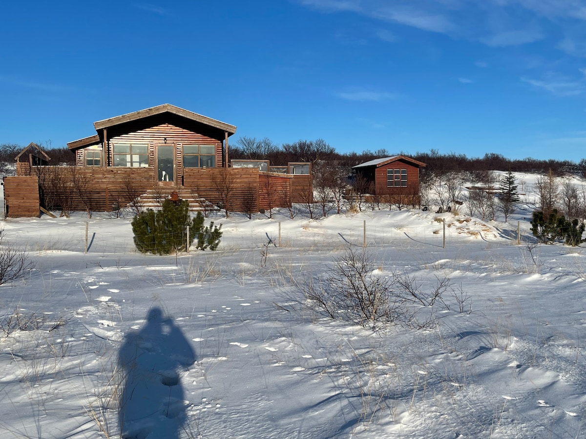 A charming wooden cabin is set against a backdrop of snow-covered land and clear blue skies. The structure features large windows that allow natural light to illuminate the interior. Nearby, a smaller building complements the main cabin, surrounded by sparse winter foliage.