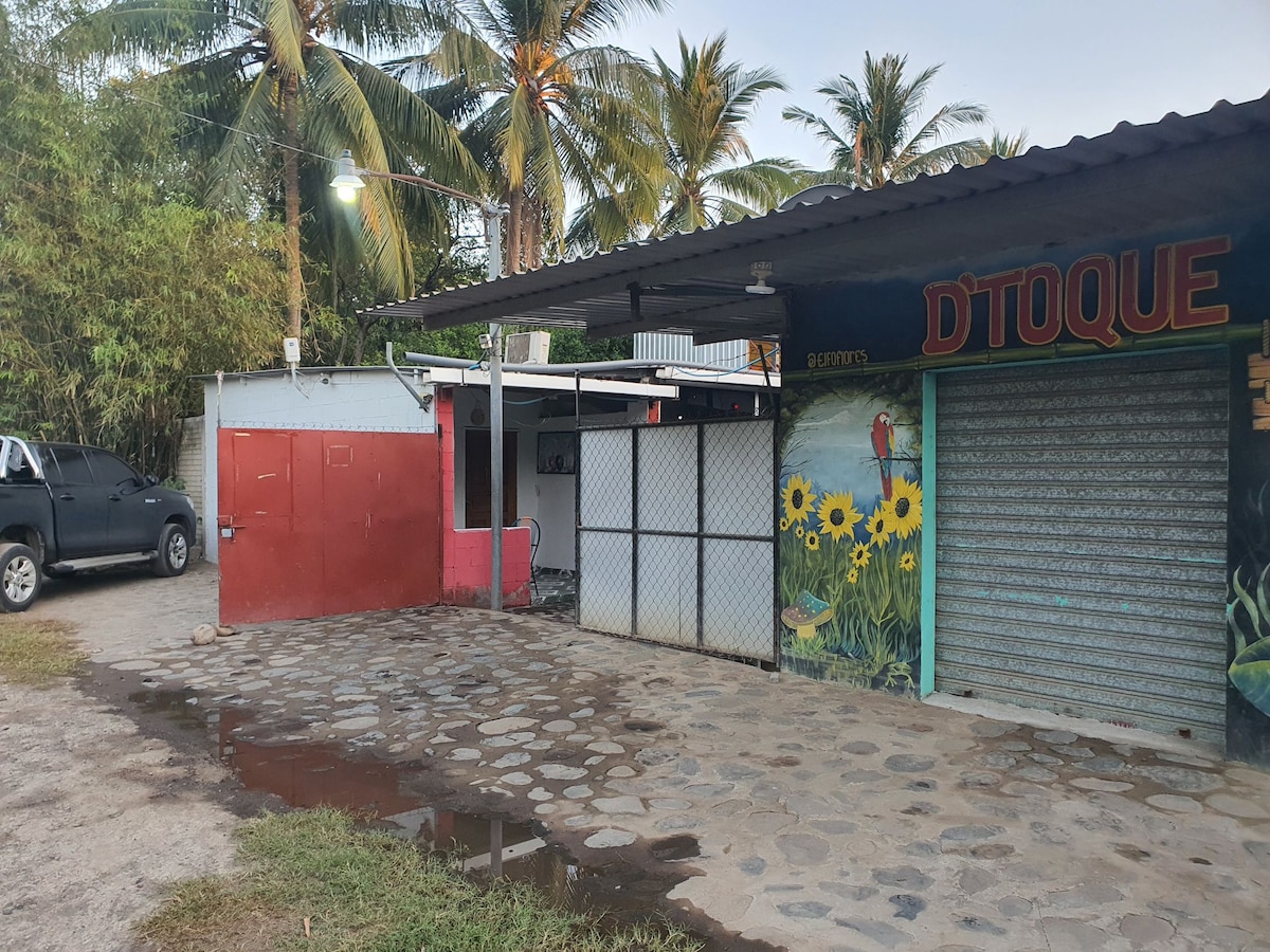 An open area features a colorful mural of sunflowers on one wall, while another wall has metal grates partially closed. Several palm trees provide a tropical backdrop, and a parked vehicle is visible on the side of the paved path, which appears slightly damp.